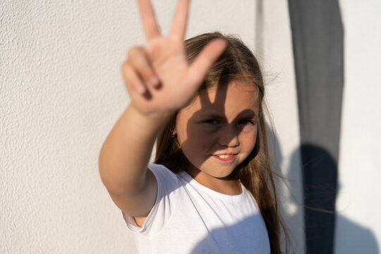 Fashion kid concept - portrait of a girl covering her face from the bright sun while standing against the white wall.
