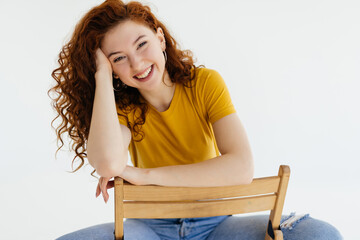 Confident charming woman in blue jeans posing on chair. Studio shot of caucasian ginger lady sitting near white wall.