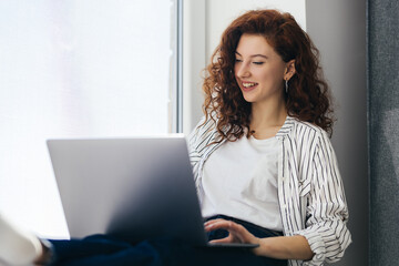 Beautiful positive woman using laptop, sitting on the windowsill in city apartment. Young red-haired woman working at home. Freelance concept.