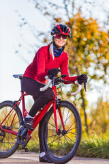 Professional Road Cycling. Winsome Caucasian Female Cyclist In Warm Outfit Posing On Road Bike While Getting Ready for Start Outdoors Against Autumn Background