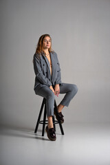 A blonde girl in a pantsuit with an unbuttoned jacket from under which can see a beautiful slender sexy body and underwear and black platform shoes poses in the studio sitting on a bar stool on white