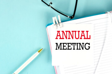 ANNUAL MEETING text on a sticky on notebook with pen and glasses , blue background