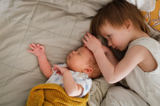 cute chubby baby sleeps on bed next to an older sibling, children are siblings together. older sister kisses and hugs the baby. In real cozy interior