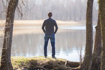 Young adult man in dark clothes standing at lake shore between trees and looking far away. Thinking...