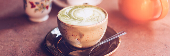 Green Matcha Latte, popular trendy tea with latte art heart shape on top. Served in cafe in ceramic mug on rustic table. Strong shadows and summer sun light