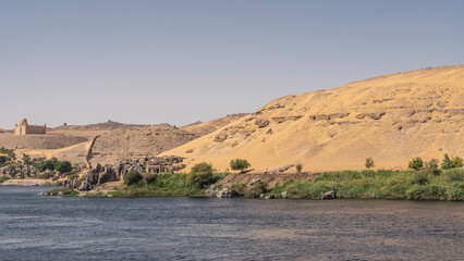 On the river bank there is a strip of green vegetation, boulders. High sand dunes against the sky. The old mausoleum of the Aga Khan is visible. Ripples on the blue water. Egypt. Nile. Aswan