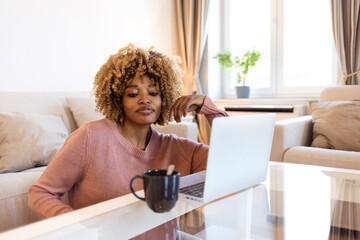 A joyful African-American young lady is sitting on the couch at home, using a laptop, watching videos online, chatting with friends or reading blogs, drink coffee. Copy space.