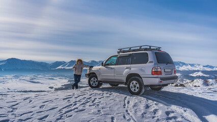 A man in a down jacket stands on the edge of a snowy plateau, leaning on an SUV. Footprints in the...