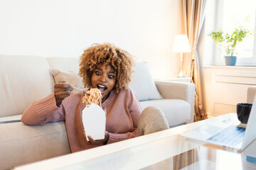 A beautiful mixed race woman eats pasta for lunch while sitting in a modern apartment at a table. The girl is busy on her laptop and that is why she uses the food delivery service.