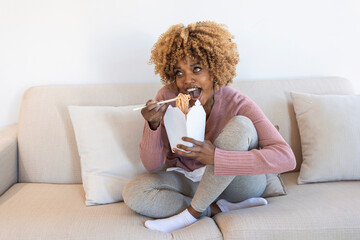 An African-American woman eats a pasta salad at home, a happy face, sits on a sofa and eats fast food that she ordered through the app. The concept of fast food and delivery