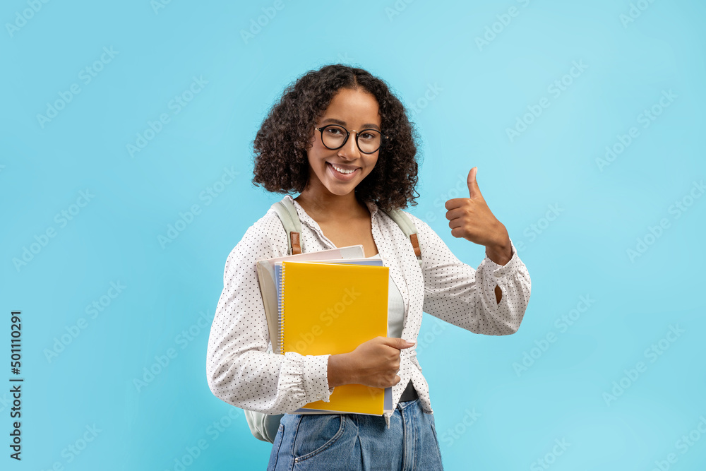 Wall mural Cheery black female student with backpack and notebooks showing thumb up, recommending modern academic education