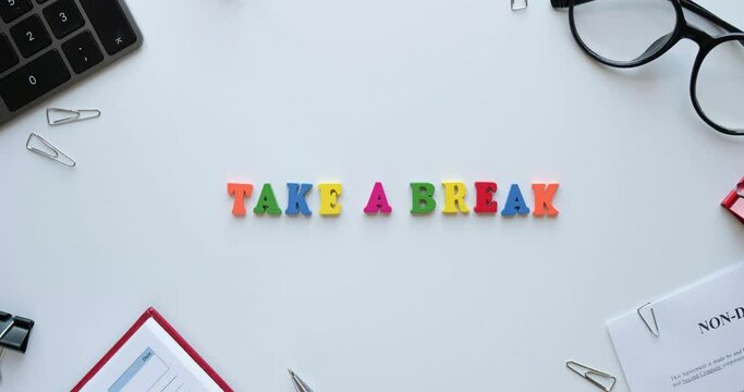 Flatlay with office items and phrase Take a break from colored wooden letters. Layout on a white background. Glasses, keyboard, pen, paper clips, diary, work papers. 4K looped stop motion animation