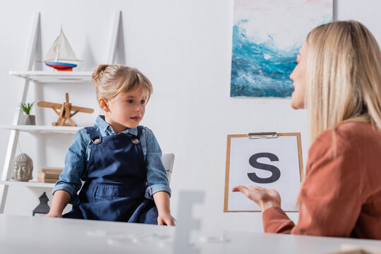 Girl sitting near blurred speech therapist with letter on clipboard in consulting room.