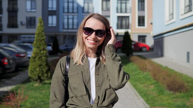 Girl wide smiling with healthy white teeth. Beautiful blonde girl adjusts sunglasses with her hand and smiles at the camera on the background of city buildings. Portrait of pretty female face model.