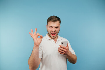 Happy adult man with smartphone in hands shows gesture ok and looks at camera with joyful emotional...