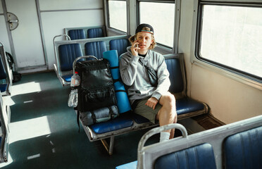 Fototapeta na wymiar Positive male tourist in casual clothes sits on a bench in a train with a backpack and talks on the phone with a smile on his face.