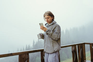 A male tourist stands in the morning on the terrace of a house in the mountains with a cup of tea in his hand and uses a smartphone.