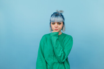 Pensive woman in green sweatshirt and with blue hair isolated on blue background with serious face.