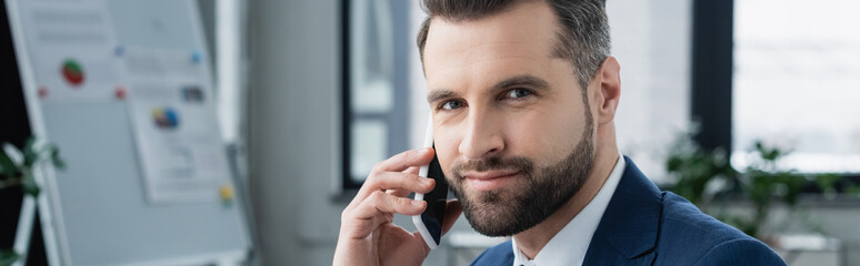 bearded businessman talking on cellphone and looking at camera in office, banner.
