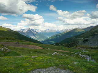 Fototapeta na wymiar Panoramic view of the beautiful landscape in the Alps with fresh green meadows and blooming flowers and snow-capped mountain tops in the background on a sunny day with blue sky and clouds in spring