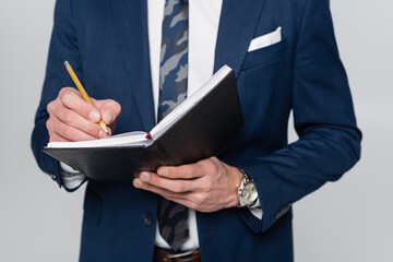 cropped view of economist in blue blazer writing in notebook isolated on grey.
