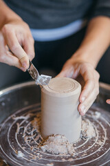 Female hands sculpt clay dishes. Сraftswoman in apron sitting at pottery wheel and using craft tool while shaping wet clay vessel