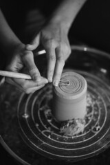 Female hands sculpt clay dishes. Сraftswoman in apron sitting at pottery wheel and using craft tool while shaping wet clay vessel