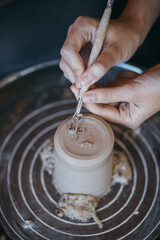 Fototapeta na wymiar Female hands sculpt clay dishes. Сraftswoman in apron sitting at pottery wheel and using craft tool while shaping wet clay vessel