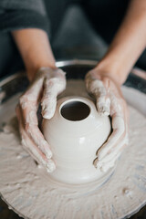 Fototapeta na wymiar Female hands sculpt clay dishes. Сraftswoman in apron sitting at pottery wheel and using craft tool while shaping wet clay vessel