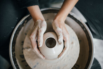 Female hands sculpt clay dishes. Сraftswoman in apron sitting at pottery wheel and using craft tool while shaping wet clay vessel