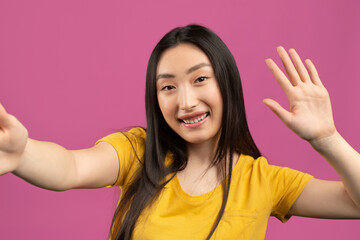 Hello. Happy korean lady taking selfie and waving hand at camera, gesturing hi while making photo over violet background