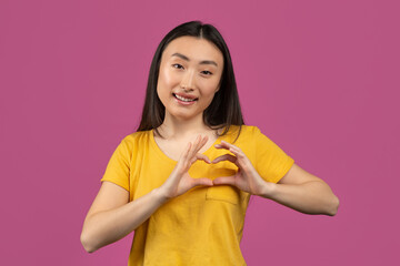 Happy asian lady showing heart gesture with hands near chest, making love sign, posing over violet...