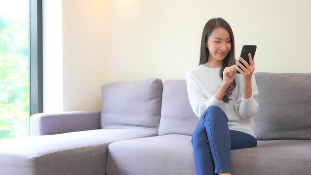 A young attractive woman in jeans and a sweater sits on a couch as she types information into her smartphone. Title space
