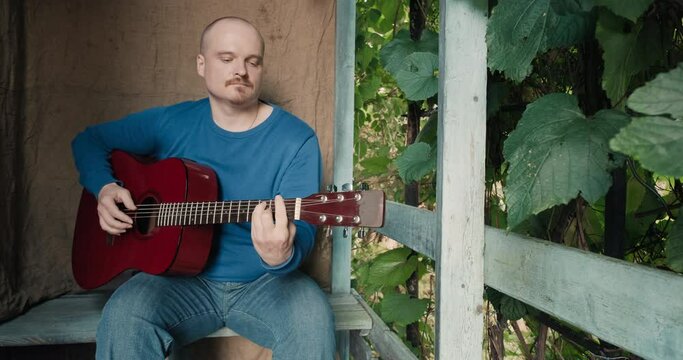 Man With An Acoustic Guitar Is Sitting On The Porch Of Farmhouse, Playing An Instrument And Singing Songs. Concept Of Creativity, Hobbies, Lifestyle And Relaxation. Old, Retro Decor, Burlap Background