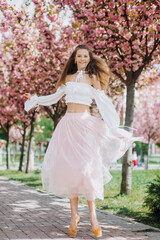 Obraz na płótnie Canvas Amazing young ballerina in an pink-white dress in pointe shoes near the pink sakura tree