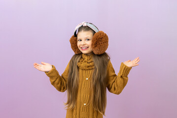 A joyful little girl in warm headphones looks at the camera, a child in a warm sweater on an isolated background.