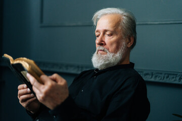 Close-up side view of senior gray-haired aged male reading paper book sitting on chair in dark living room, relax and rest with literature. Handsome mature adult man reading book at home alone.