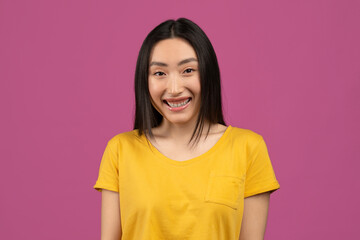 Portrait of cute asian woman in yellow t-shirt posing over violet studio background, looking at...