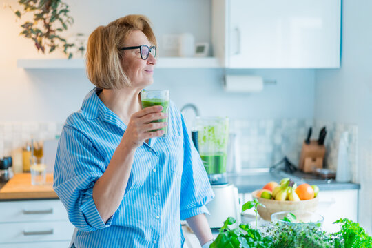 Smiling middle aged woman with just made glass of detox shake, green smoothie in the kitchen. Healthy dieting, eating, cooking. Natural anti aging methods, weight loss program. Vegan, vegetarian diet