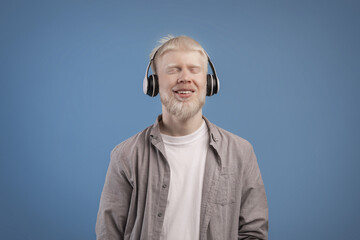 Satisfied calm albino guy in headphones enjoying favorite music with closed eyes standing over blue background