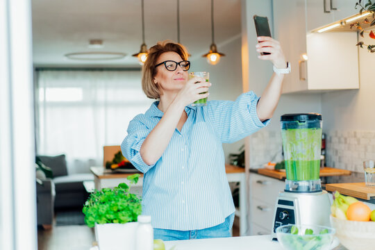 Middle aged woman making selfie with just made glass of detox shake, green smoothie in the kitchen. Healthy dieting, eating, cooking, weight loss program. Blogger creating content for social media.