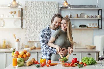 Cheerful young caucasian lady cooking salad, guy with stubble hugs woman at table with organic vegetables