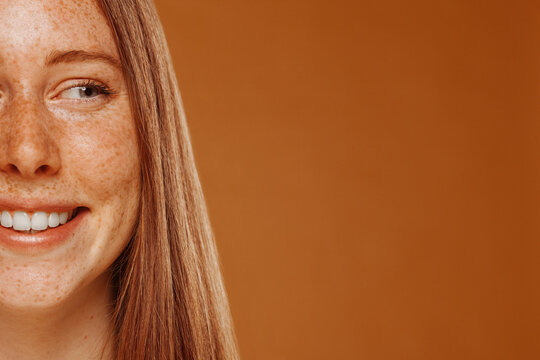 Half face beauty portrait of slim redhead beautiful happy smiling model with freckles and ginger hair, standing over orange background, looking away. Copy space. Cosmetic, skincare concept.