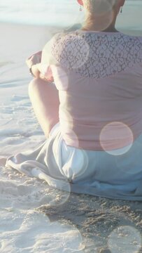 Animation of light spots over senior caucasian woman practicing yoga at beach and meditating