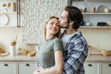 Happy millennial european husband with stubble hugs wife behind, kisses at forehead in kitchen...