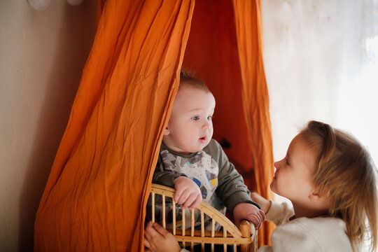 cute baby in wicker baby bed and under canopy of terracotta color and an older sister. baby is in crib, sister hugs and kisses the baby in cow.