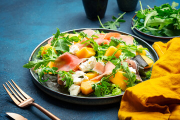 Delicious summer fresh salad with cantaloupe melon, prosciutto, soft cheese and arugula on blue table background, top view, copy space
