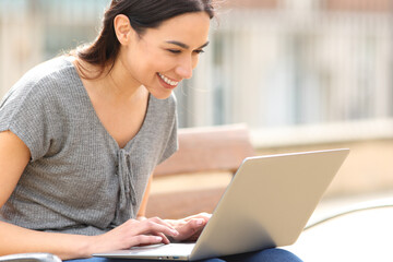 Happy woman using laptop in the street on a bench