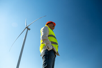 Wind turbine generates clean electric energy. Engineer in visibility vest and orange helmet stands...