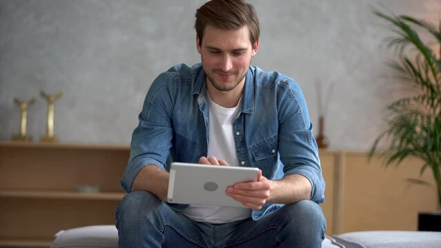 Attractive man sit on sofa holding tablet device. Do telecommute job remotely, chatting in social media, choose goods on web stores. E-commerce, virtual services, fun app user concept